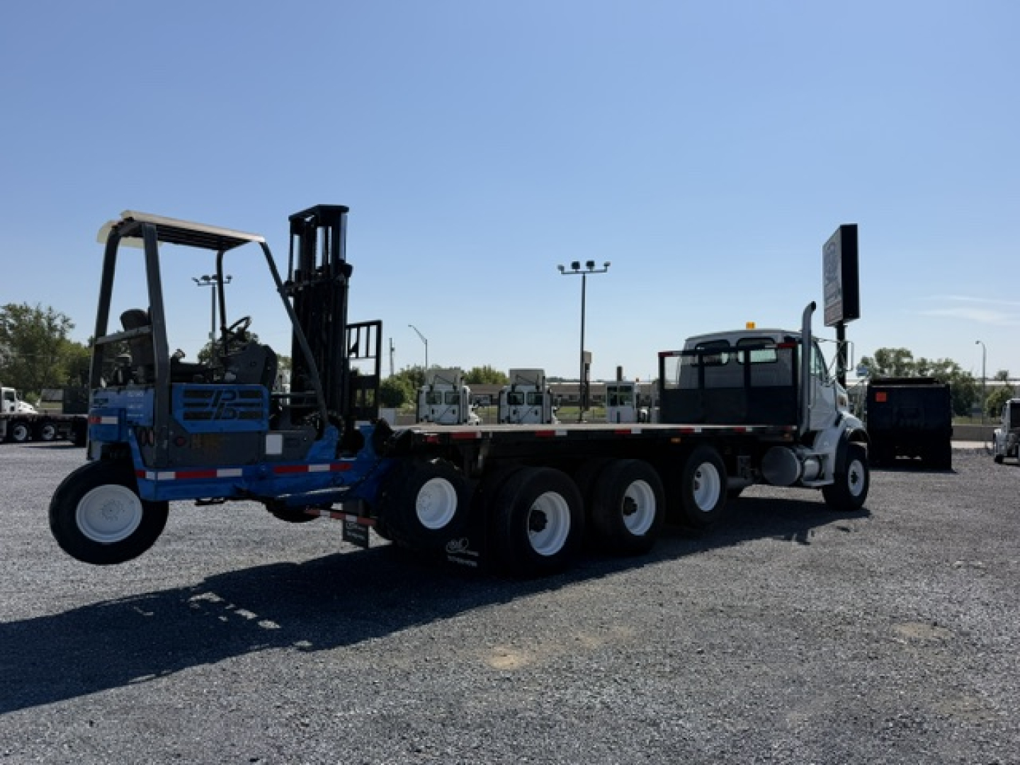 Flatbed truck tractor with a blue and black forklift attachment on a long multi-axle trailer connected to a white cab truck, in an open lot under a clear sky.