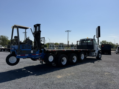 Flatbed truck tractor with a blue and black forklift attachment on a long multi-axle trailer connected to a white cab truck, in an open lot under a clear sky.