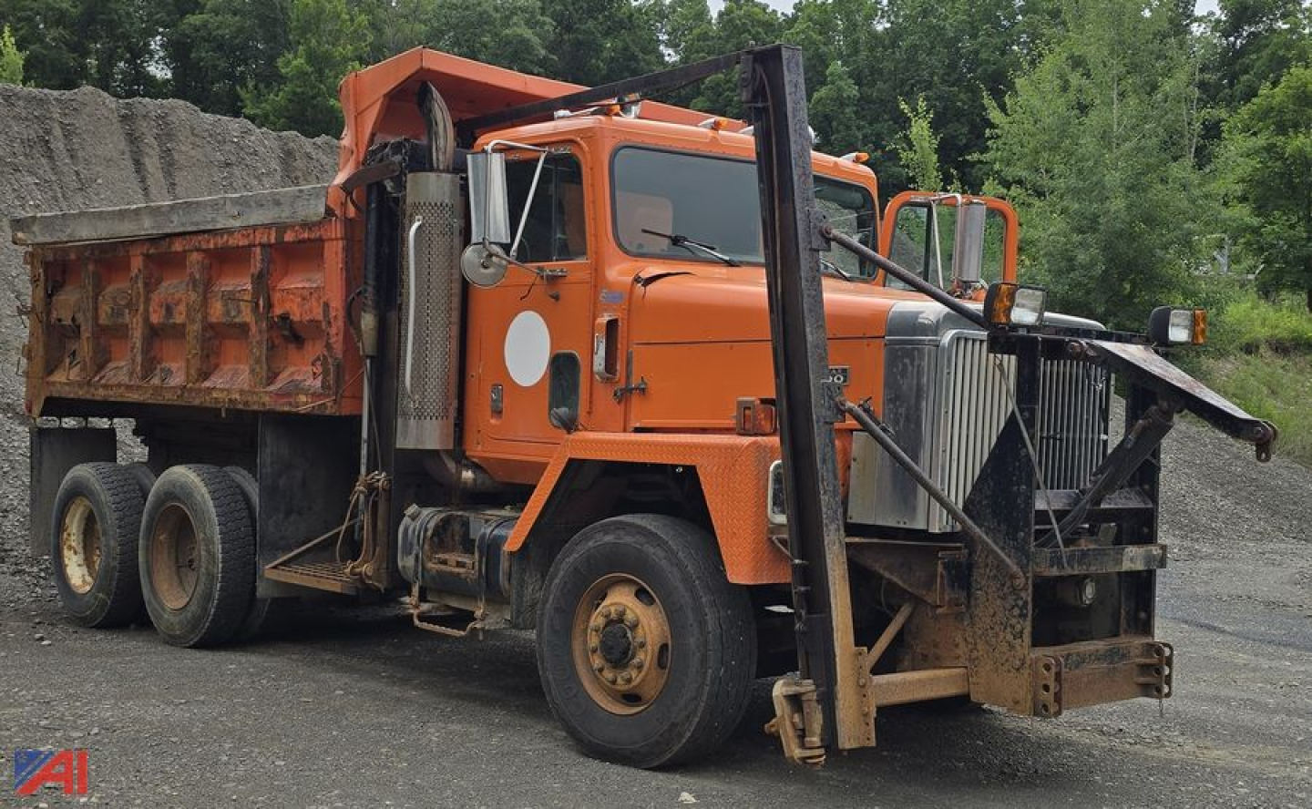 Dump truck with orange cab and dump bed showing front bumper, stacked dual rear axles, and heavy-duty suspension in an outdoor yard.