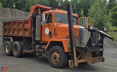 Dump truck with orange cab and dump bed showing front bumper, stacked dual rear axles, and heavy-duty suspension in an outdoor yard.