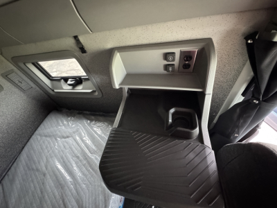 Truck interior cab area showing a storage tray and power outlet panel in a Volvo VNL sleeper cab, with window and seat nearby.
