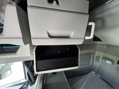 Volvo VNL truck sleeper cab interior showing overhead bunk area and storage compartment with open rectangular console, gray panels, and side window reflection.