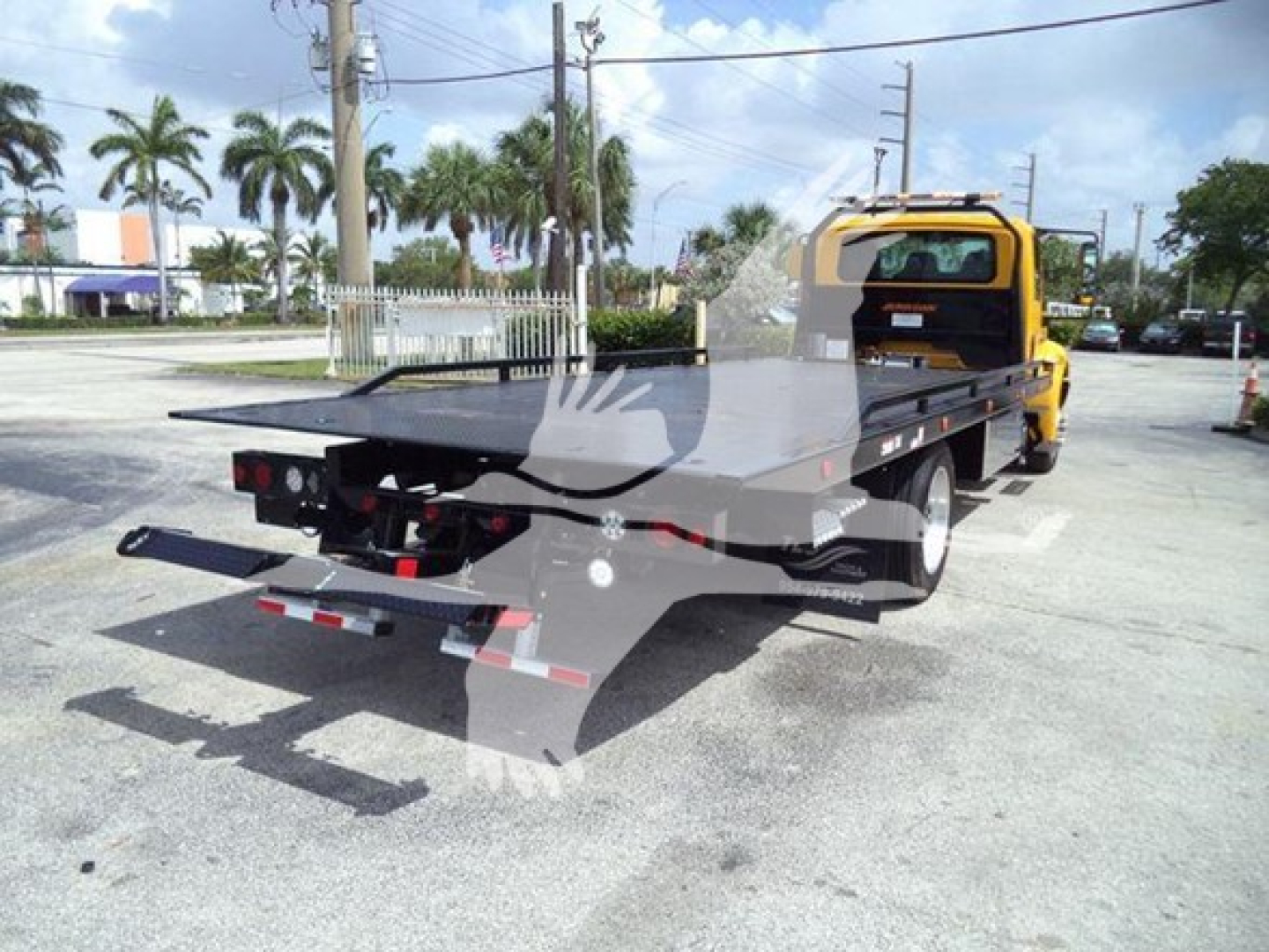 Flatbed tow truck with yellow cab and black bed, viewed from rear-left side; license plate area not visible; rear lights and reflective tape visible.