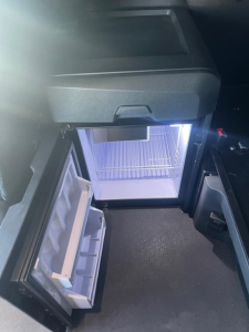 Cargo area of a Freightliner Cascadia truck interior showing an open mini fridge/freezer drawer and small refrigerator with illuminated interior.