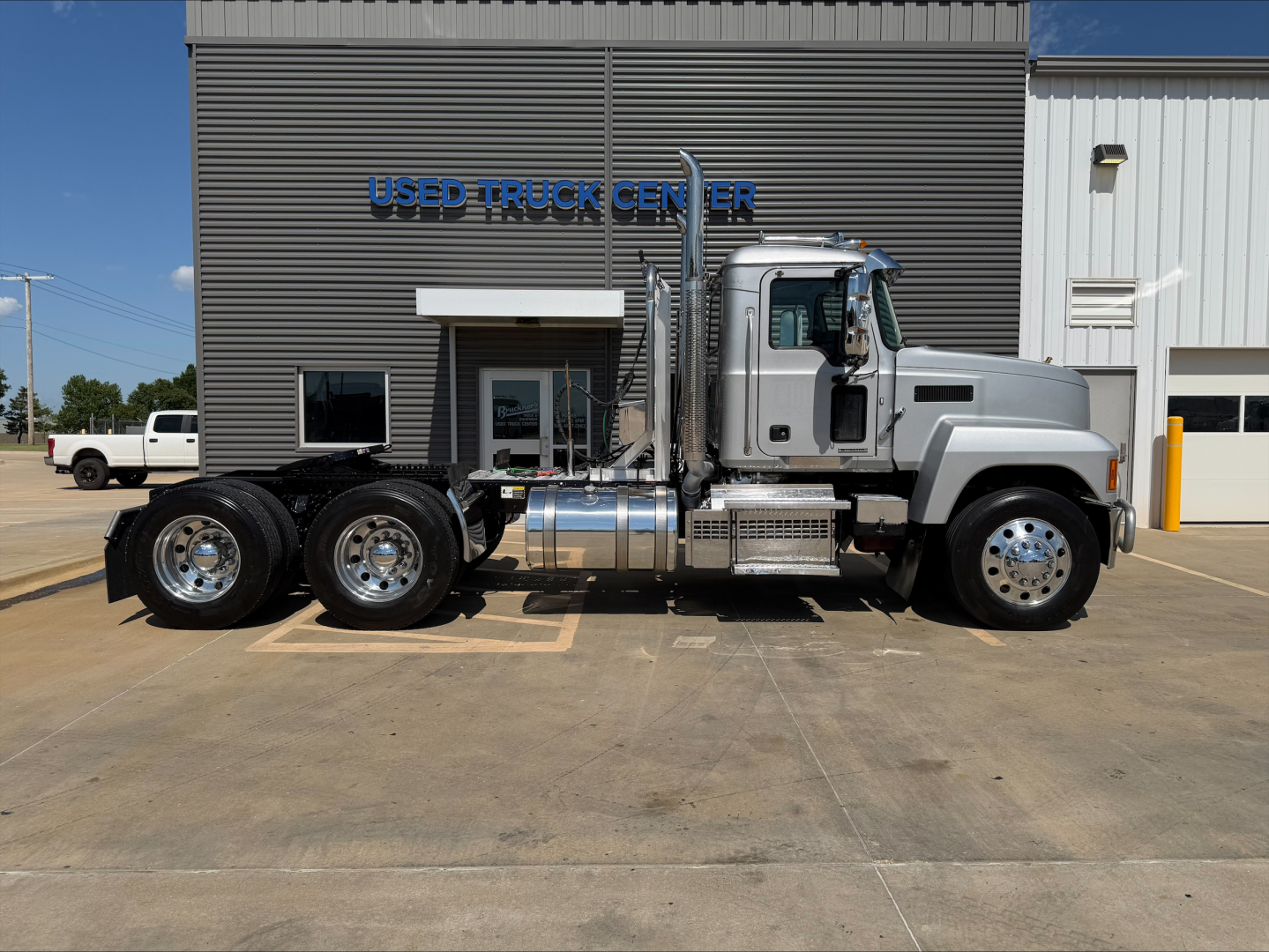 2019 Mack PI64T conventional daycab truck in side profile view assembled with dual rear axles and chrome fuel tanks in front of a dealership building.