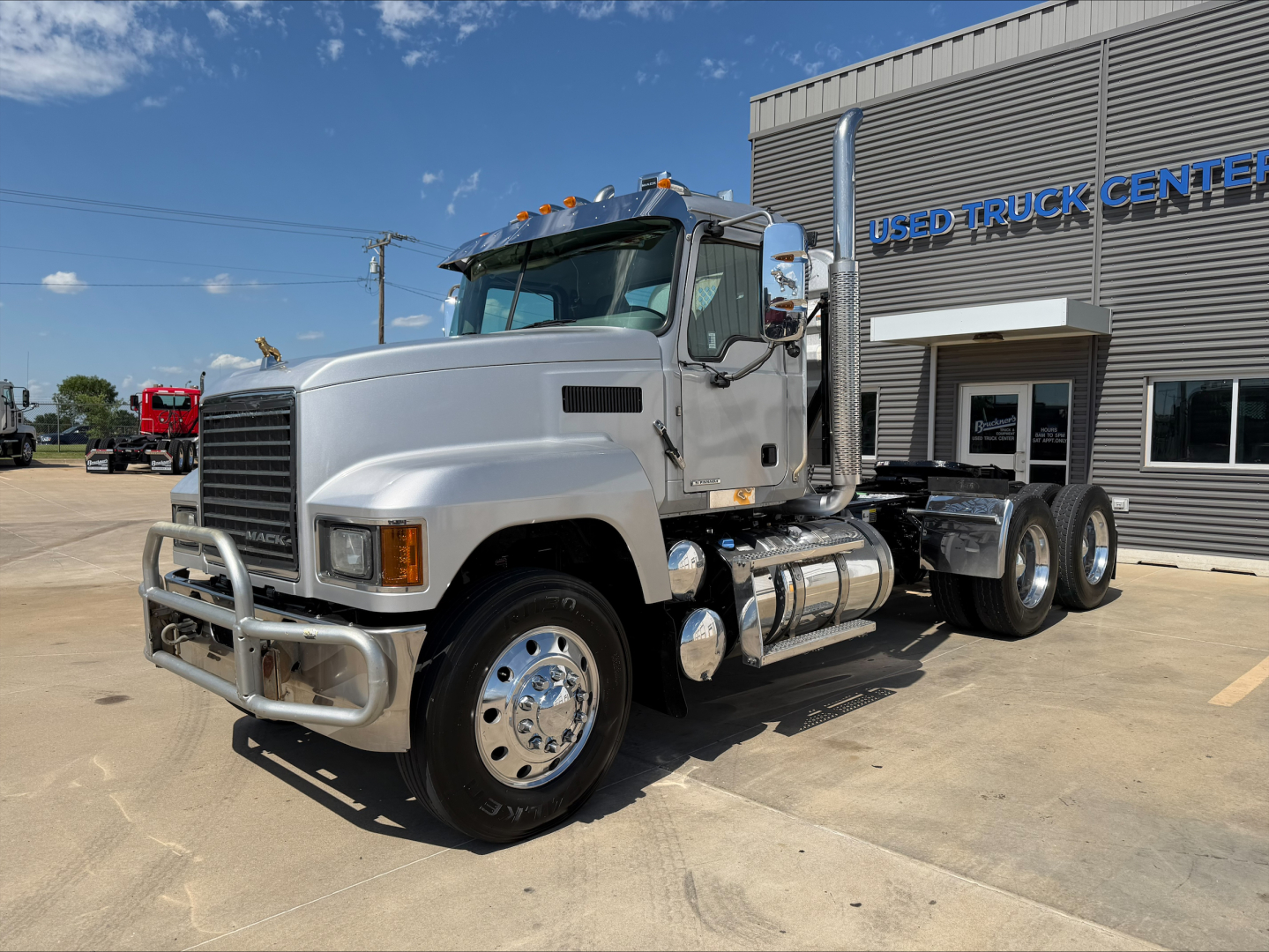 Truck chassis with cab in front, chrome bumper guard, large grille, Mack PI64T conventional daycab. Dual rear axles, aluminum fuel tanks, exhaust stack, and 'USED TRUCK CENTER' building in background.