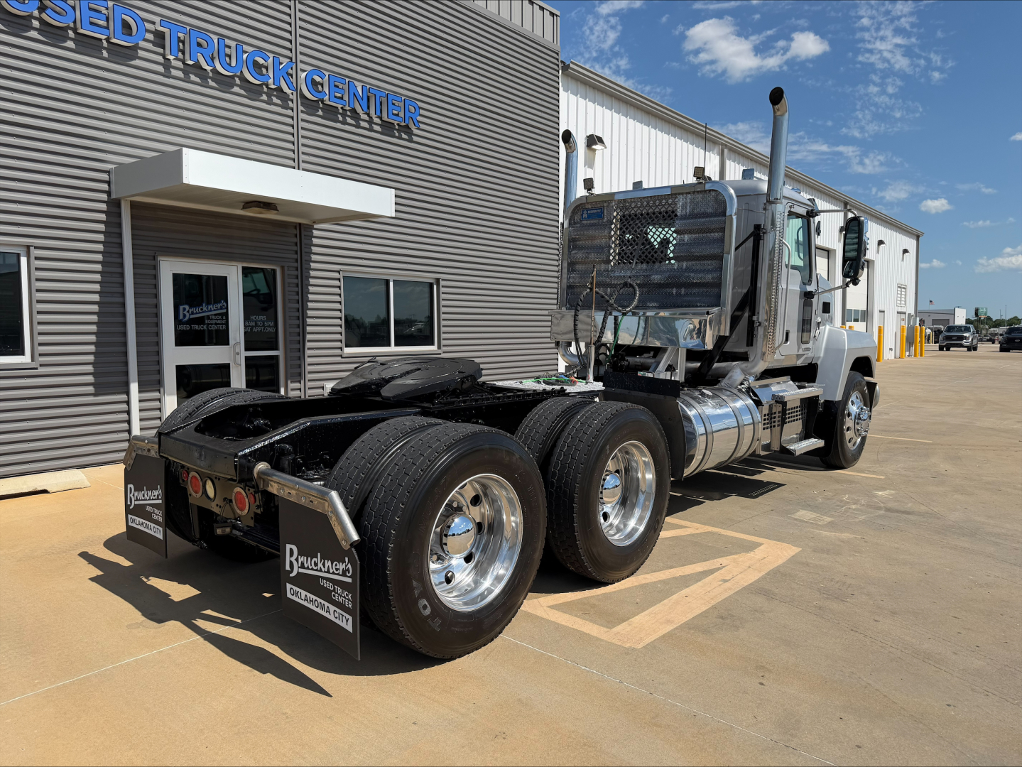Mack PI64T conventional daycab truck seen from rear-left side, dual rear axles, silver exhaust stacks, chrome fuel tanks, and shiny wheels in front of dealership building.