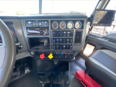 Mack PI64T cab interior showing dashboard gauges, controls, and a mounted tablet on the right; transmission shift buttons and colorful knobs below dash.