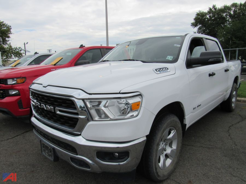 White 2023 Ram 1500 pickup truck in a dealership lot, front three-quarter view showing chrome grille, headlights, and badge; others vehicles parked nearby.