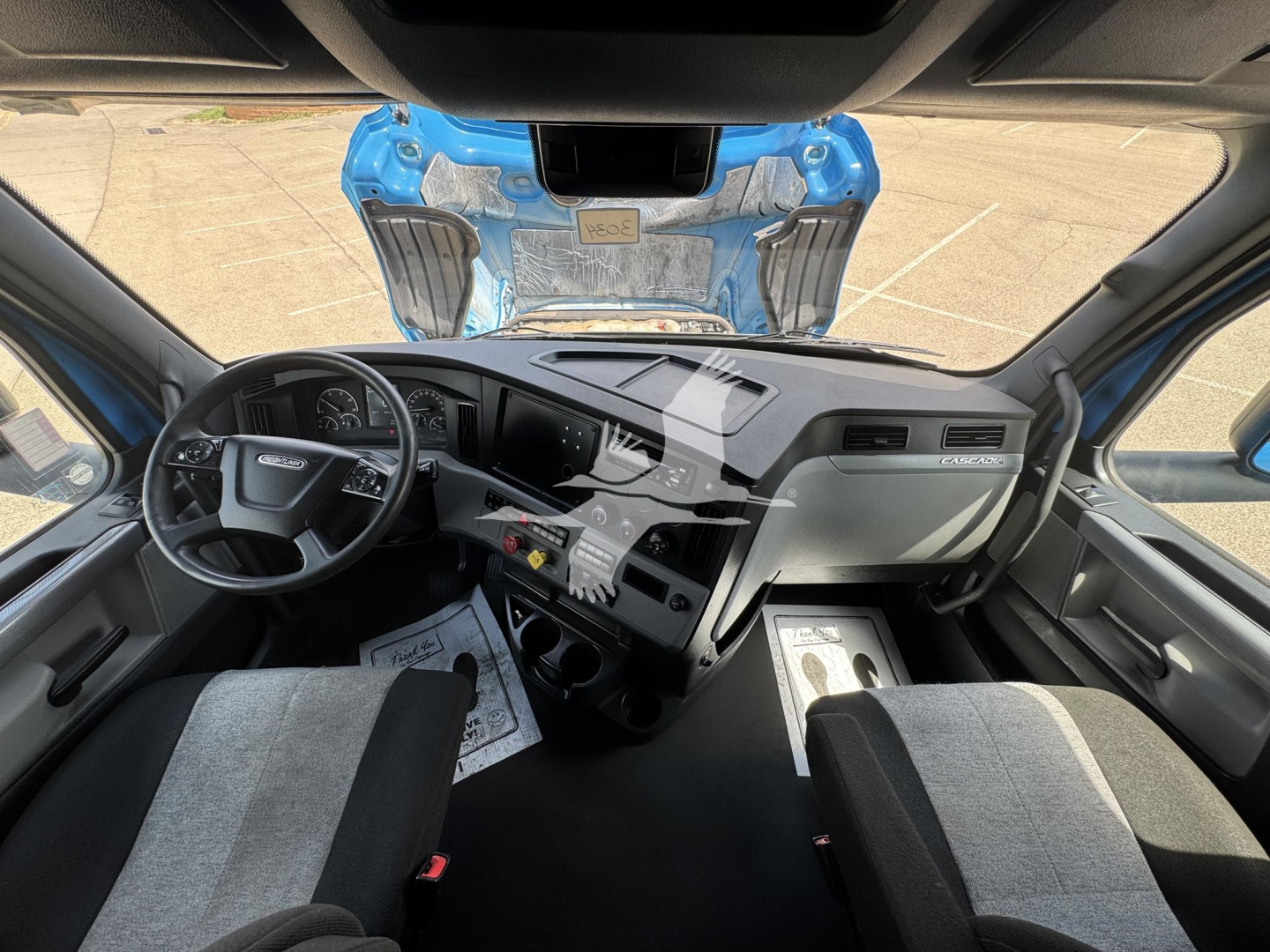 Freightliner Cascadia truck cab interior view showing steering wheel, dashboard, and center console; visible overhead bunk mechanism outside windshield.