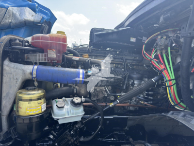 Truck engine bay of a 2023 Freightliner Cascadia 126 showing coolant reservoir, hydraulic fluid tank, multiple colored wiring harnesses, and various hoses.