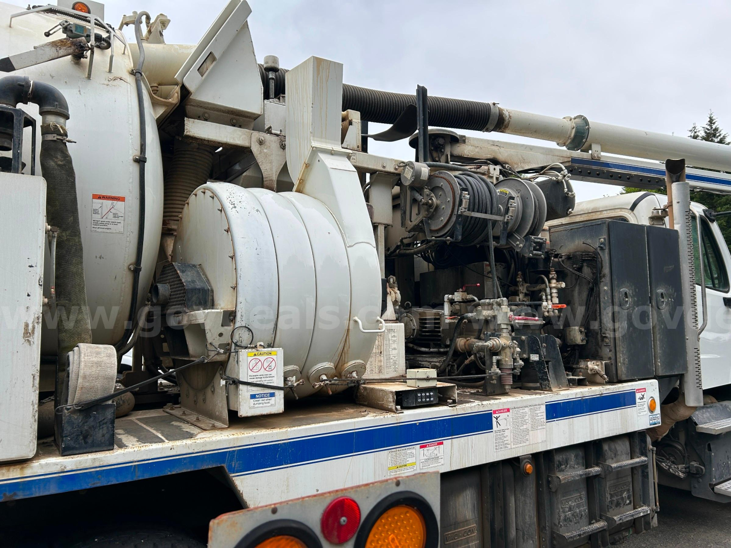 Freightliner truck engine bay showing large vacuum system and hydraulic components on flatbed with white cylindrical tanks and hoses.