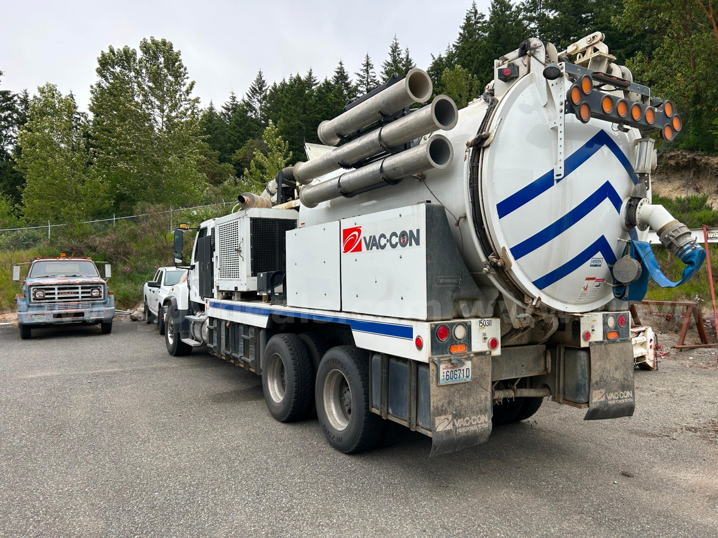 Truck-mounted vacuum tanker with VacCon logo, multiple hoses coiled atop, rear hatch, and dual rear axles. Side text shows VacCon branding and a large circular rear door.