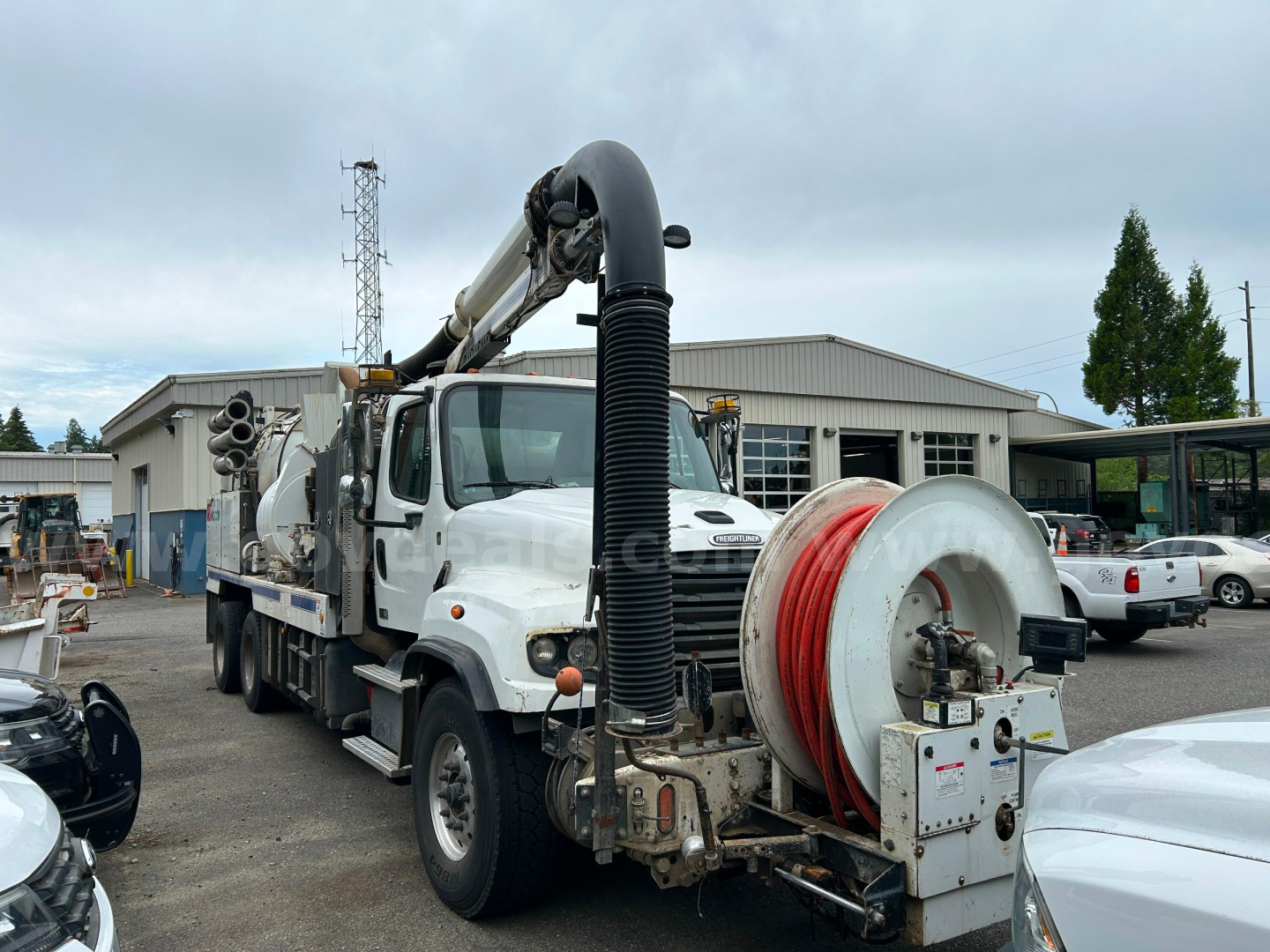 Truck body with a long vacuum hose reel and mounted hose reel on a white Freightliner unit in a yard.
