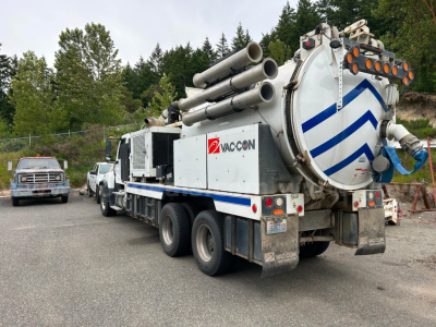 Truck-mounted vacuum tanker with VacCon logo, multiple hoses coiled atop, rear hatch, and dual rear axles. Side text shows VacCon branding and a large circular rear door.