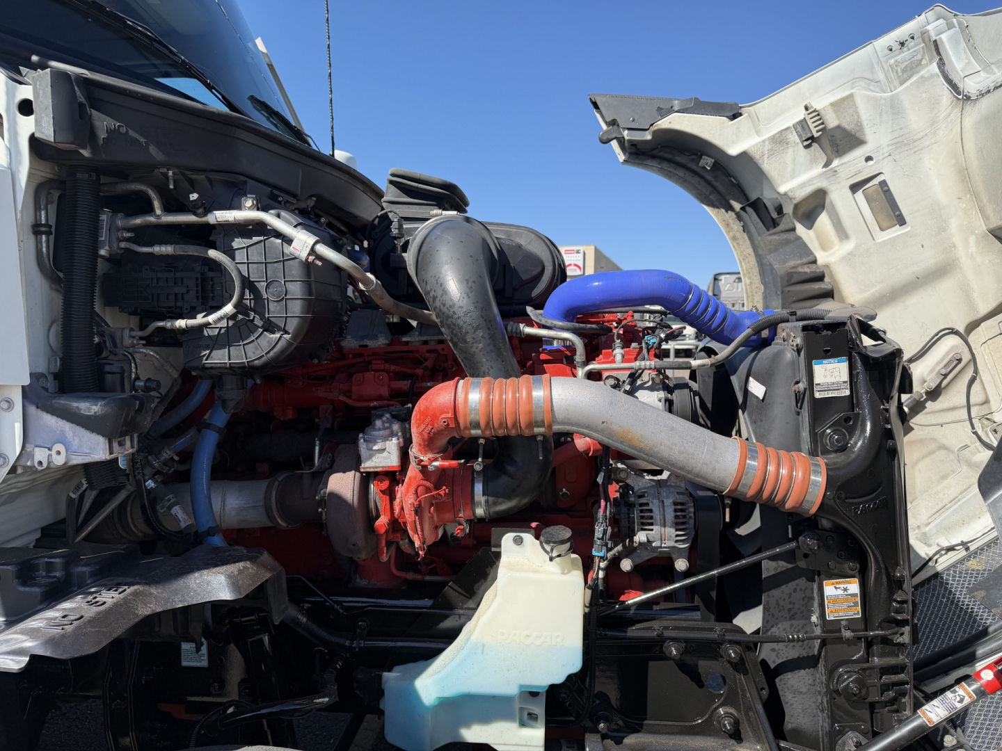 Kenworth T680 engine bay showing red diesel engine, black hoses, blue cold-side hose, orange coolant hose, and translucent coolant reservoir with light blue fluid.