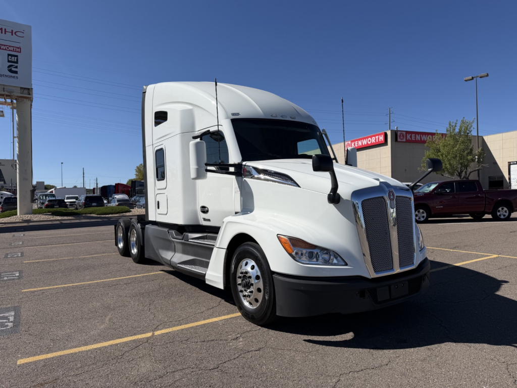 Kenworth T680 tractor-trailer in a dealership lot, white exterior, long nose, sleeper cab, dual rear axles, front grille with Kenworth emblem, side mirrors, chrome wheel covers.