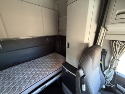 Interior of a Kenworth T680 sleeper cab showing a lower bunk bed with quilted mattress and a partition wall, passenger seat, and window curtains.