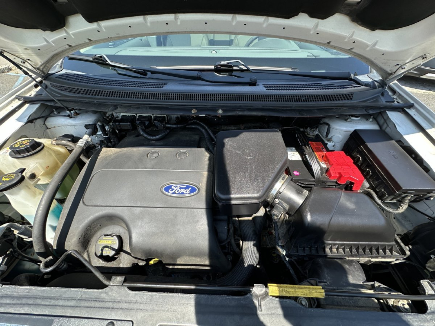 Ford engine bay showing 2.0L EcoBoost? No, image shows Ford engine with large plastic intake cover and red battery terminal cover. Battery, air filter box, coolant reservoir visible.