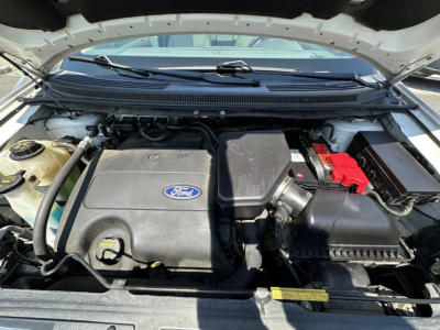 Ford engine bay showing 2.0L EcoBoost? No, image shows Ford engine with large plastic intake cover and red battery terminal cover. Battery, air filter box, coolant reservoir visible.