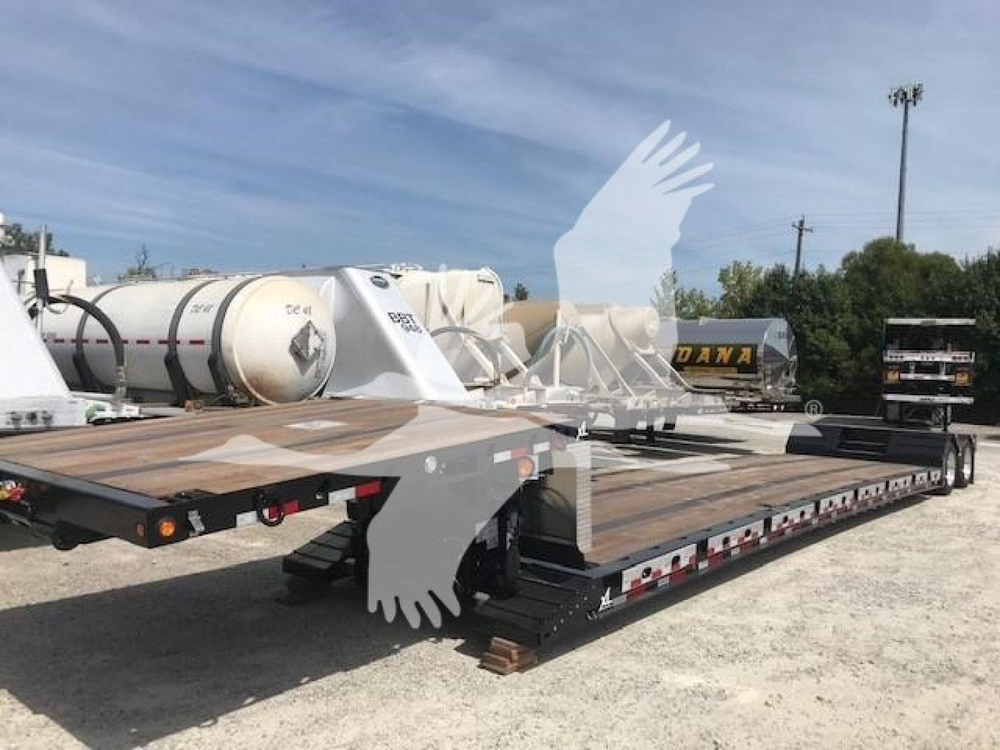 Lowboy trailer with wooden deck and metal frame, view of rear and side, with multiple white cylindrical tanks and equipment in background.