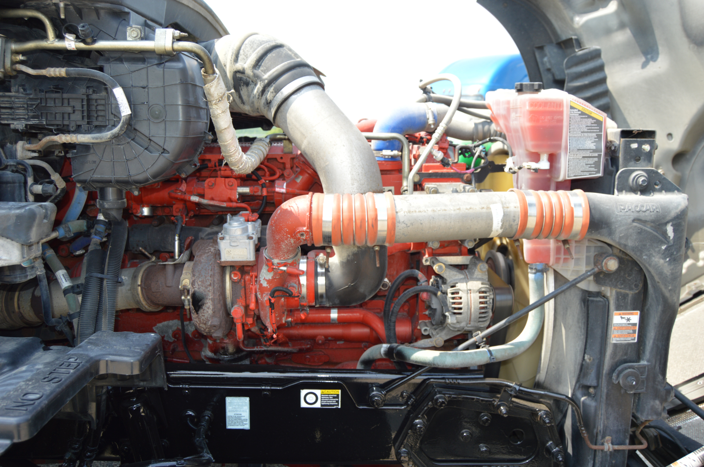 Kenworth T680 engine bay with red engine, coolant reservoir, hoses, and blower; visible components and wiring in a truck engine compartment.