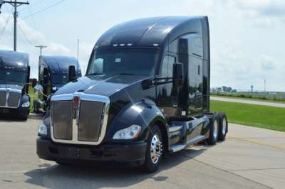 Kenworth T680 tractor in black at a dealership lot with two other trucks in background, front grille, headlights, hood and cab visible.