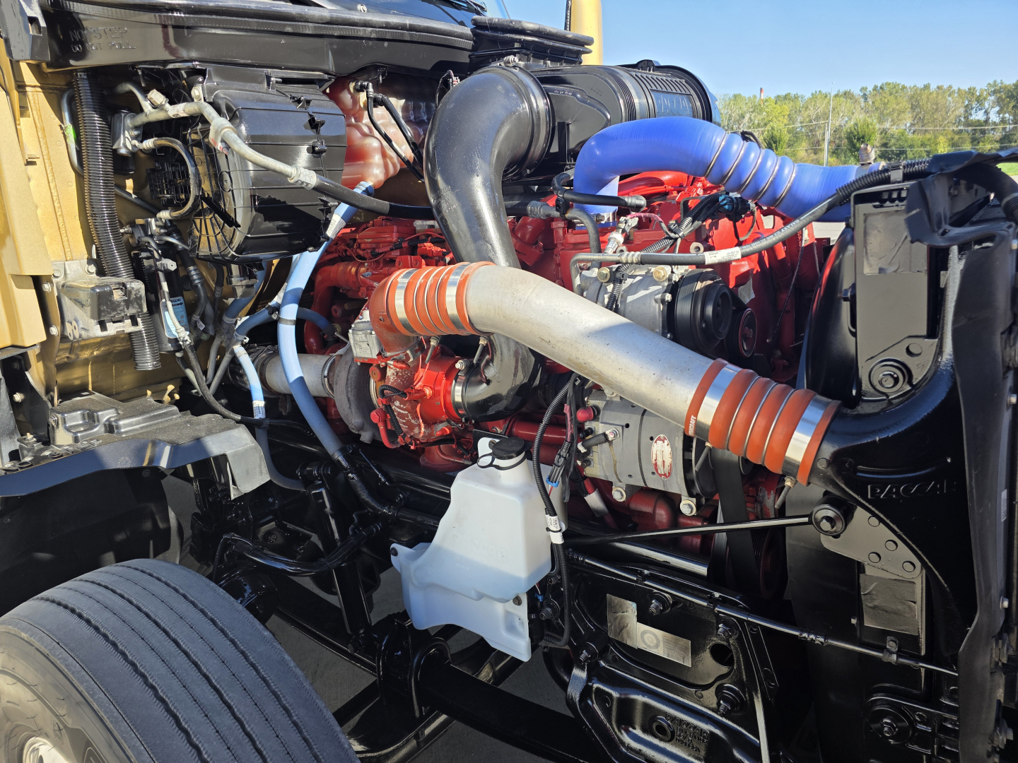 Kenworth T680 engine bay showing large turbocharger and red engine components with blue silicone hose, black frame rails and white coolant reservoir.