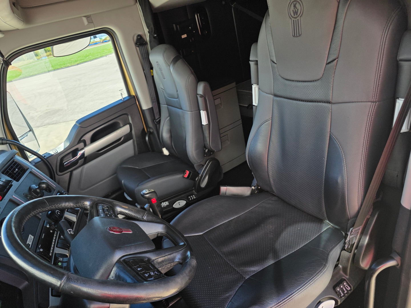 Kenworth T680 truck interior showing driver seat and passenger seat, steering wheel with Kenworth emblem, dashboard, and cab controls.