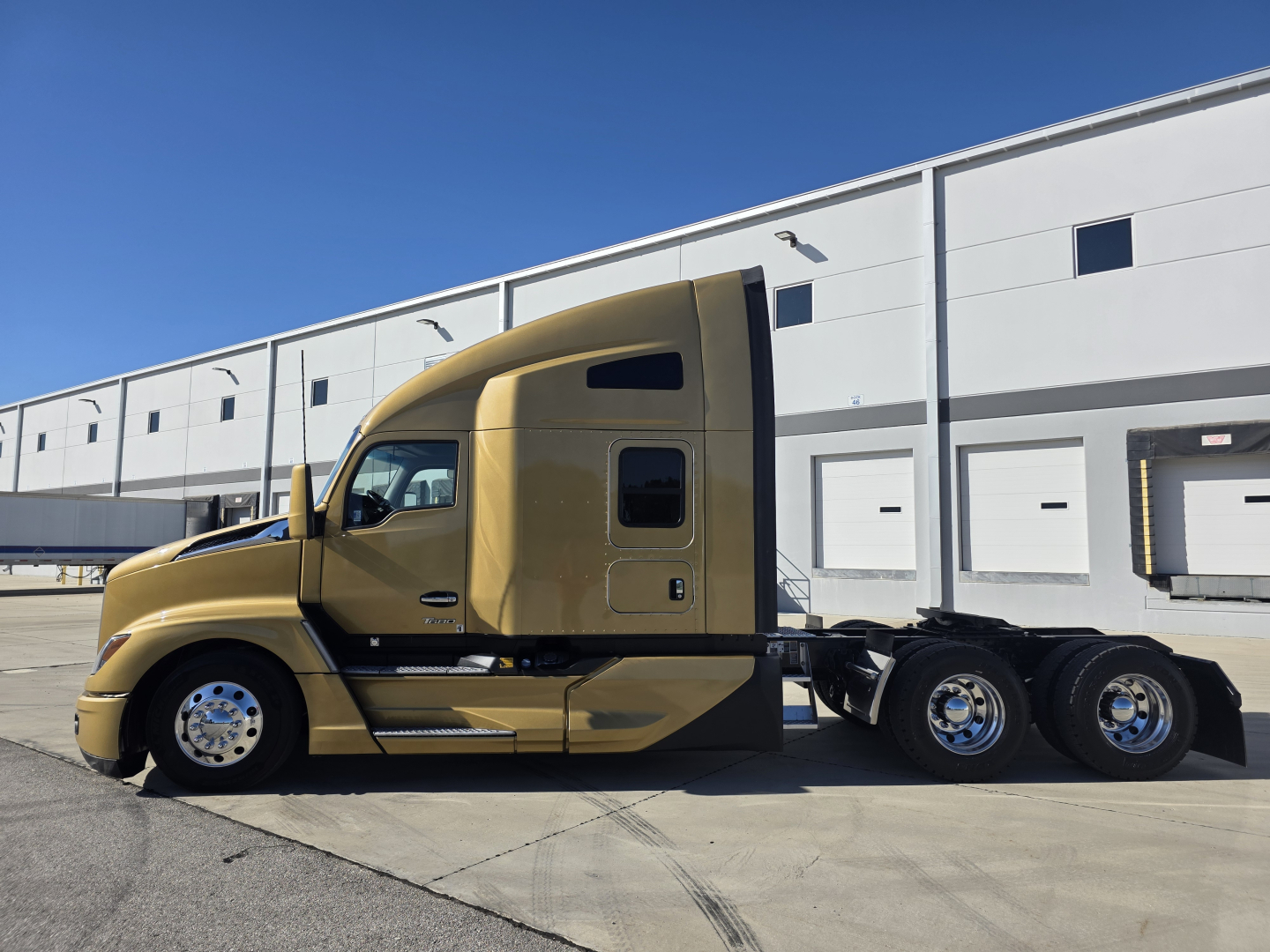 Kenworth T680 sleeper cab tractor in tan color, side profile view, attached to a long trailer, parked outside a warehouse.