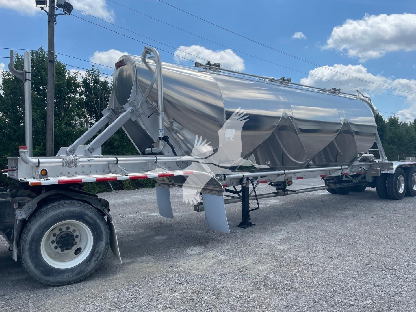 Pneumatic dry bulk trailer in polished metal on a gravel lot, attached to a truck tractor; side view shows large cylindrical tank with support frame and wheels.