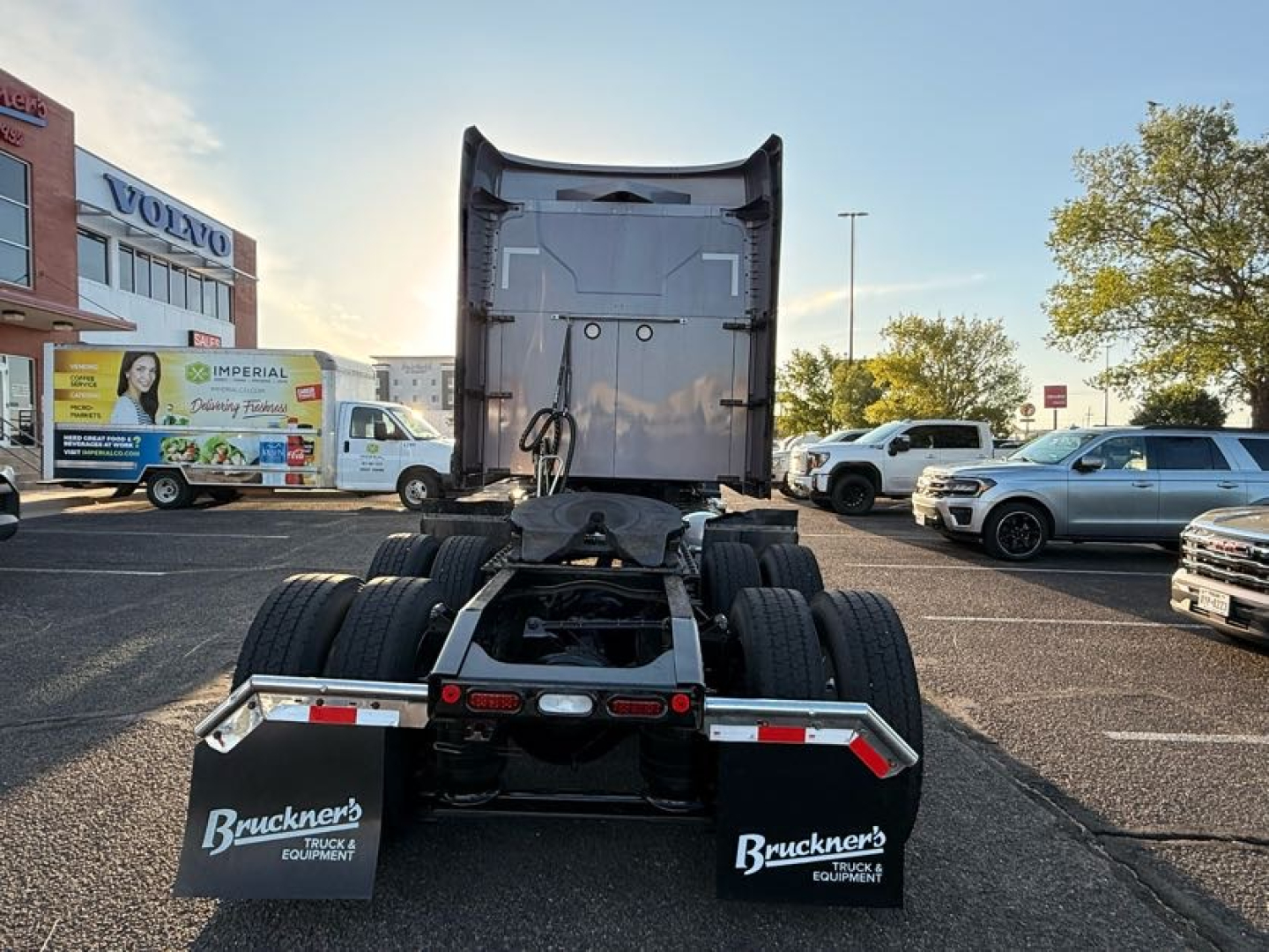 Truck rear view showing fifth wheel and dual rear axles with mud flaps; visible text on mud flaps reads 'Bruckner's TRUCKS EQUIPMENT' and license plate area; surrounding vehicles in lot.