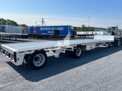 Flatbed trailer in a gravel lot with a white aluminum deck and dual rear axles, a Sherwin-Williams blue trailer on another lot in the background.
