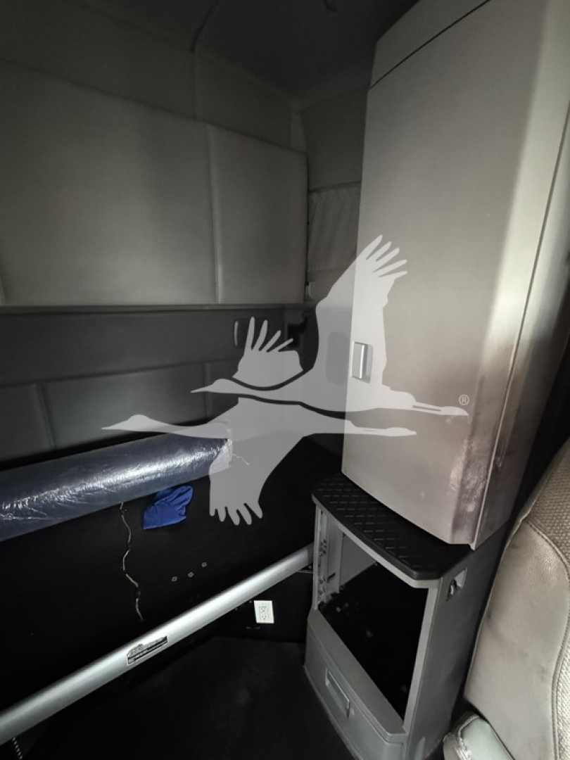 Cabin interior of a Kenworth truck showing a storage compartment and a small open shelf with a drawer. The image includes a vertical cabinet, a metal bar/pole, and a seat edge visible.