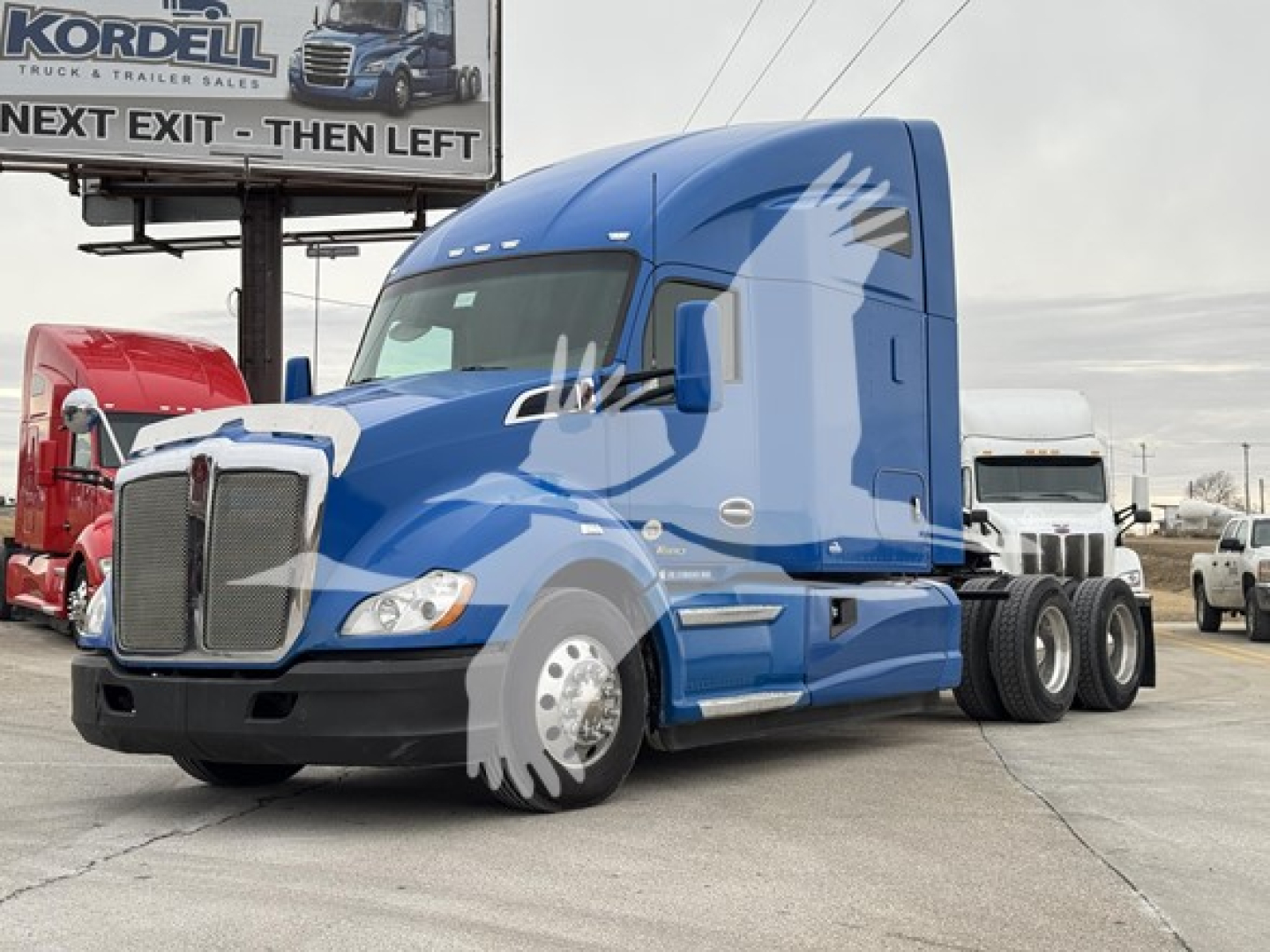 Kenworth T680 tractor-trailer in blue, side view, with stock and sales lot background.