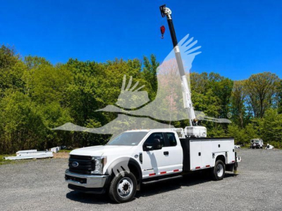 Ford F-550 service/utilities truck with bucket lift, white cab and utility bed, in a gravel lot with trees in background.