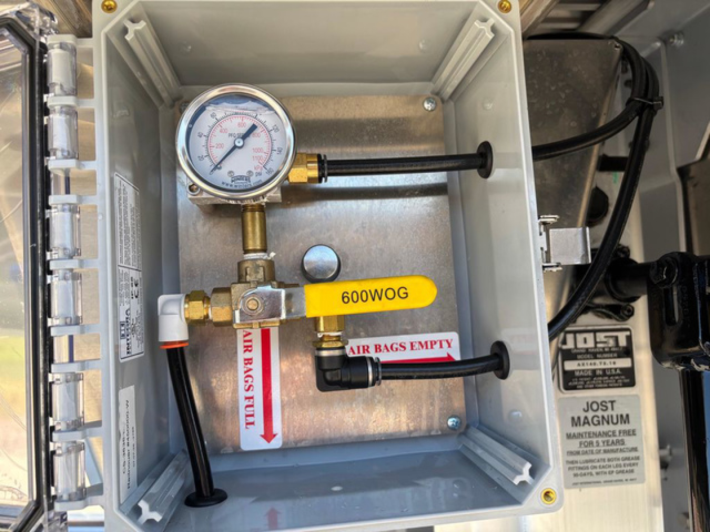 Dropdeck trailer reservoir panel with pressure gauge and label 600WOG. Visible features: pressure gauge, brass valve, black hoses, yellow tag 600WOG, white label AIR BAGS FILL, metal box housing, JOST Magnum label on adjacent component.