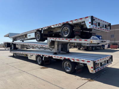 Dropdeck trailer stacked two levels showing aluminum flatbed sections, wheels, reflective safety tape, and spare tire, Reitnouer brand with dual axles and ramp sections.
