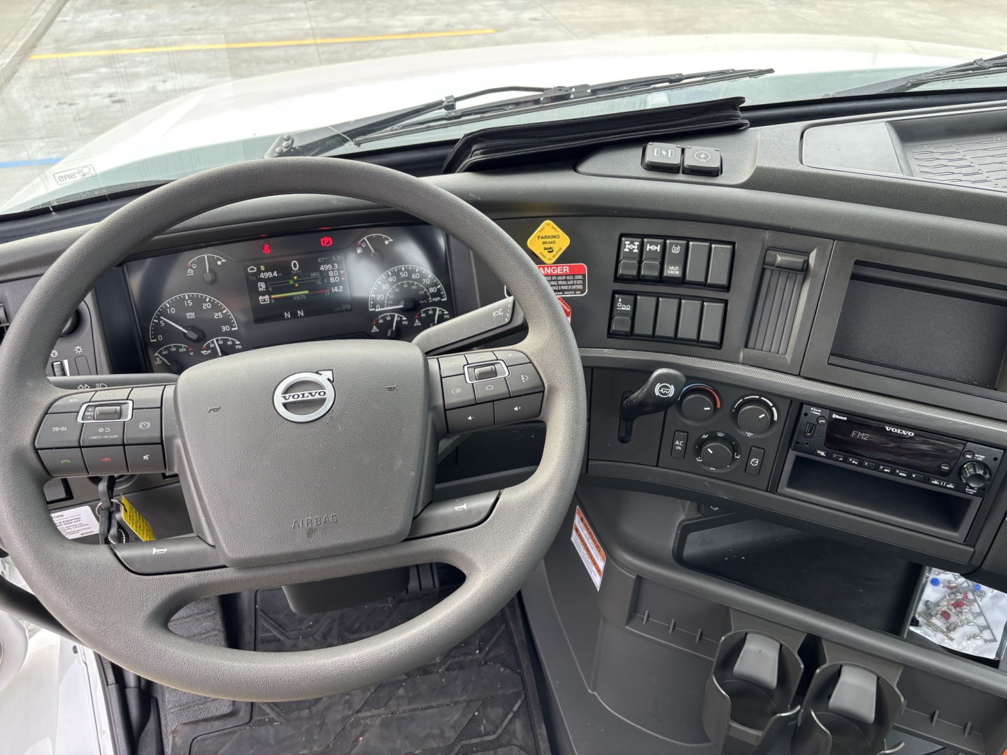 Volvo truck cockpit with steering wheel and dashboard, multiple control switches, circular gauges, and center console; visible labels include Volvo brand emblem on steering wheel.