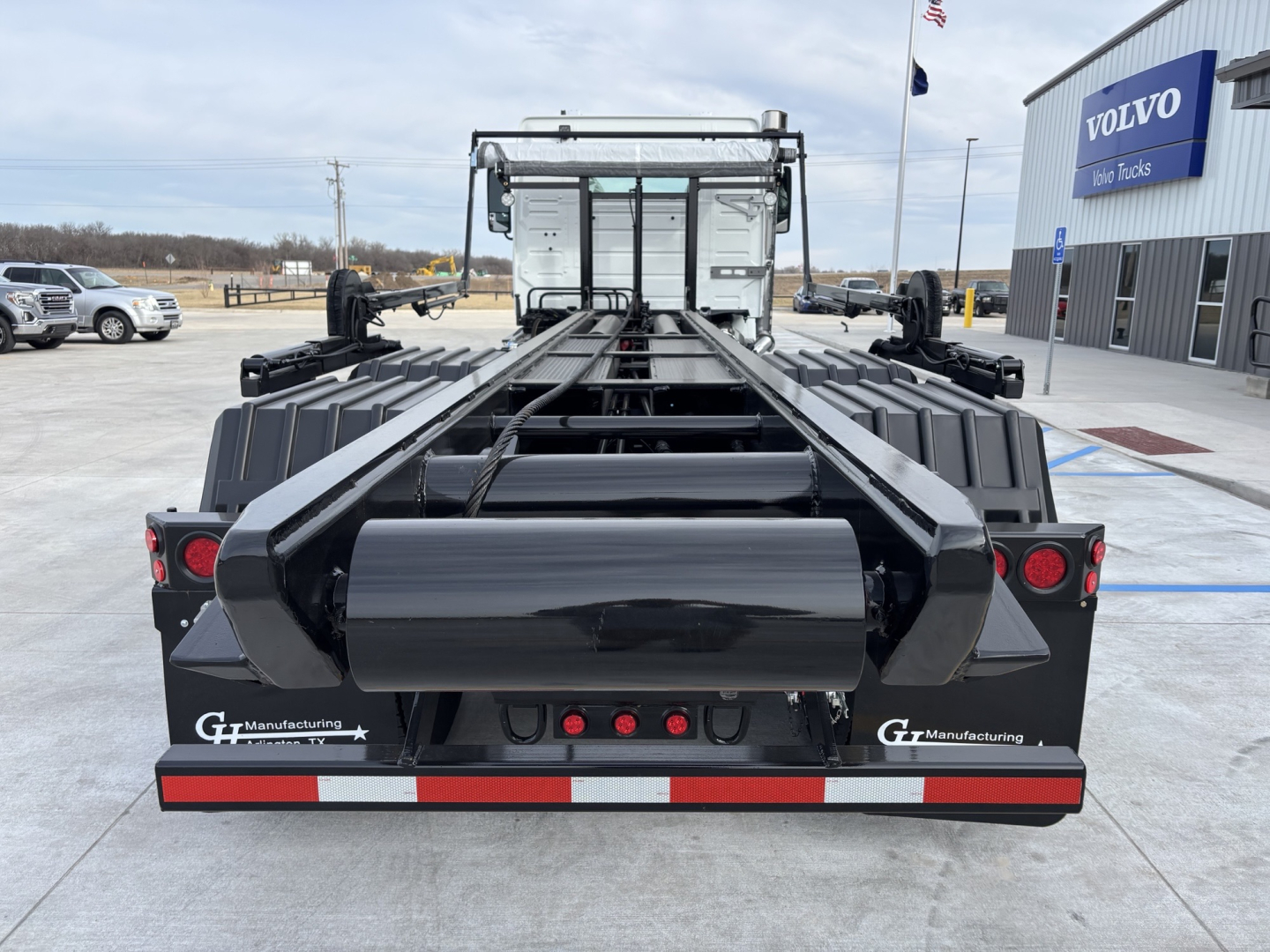 Rear view of a Volvo VHD64B300 roll-off truck chassis showing black flatbed with bed arms and tail lights; visible branding reads "G7 Manufacturing" on both sides and reflective red/white stripe at bumper.