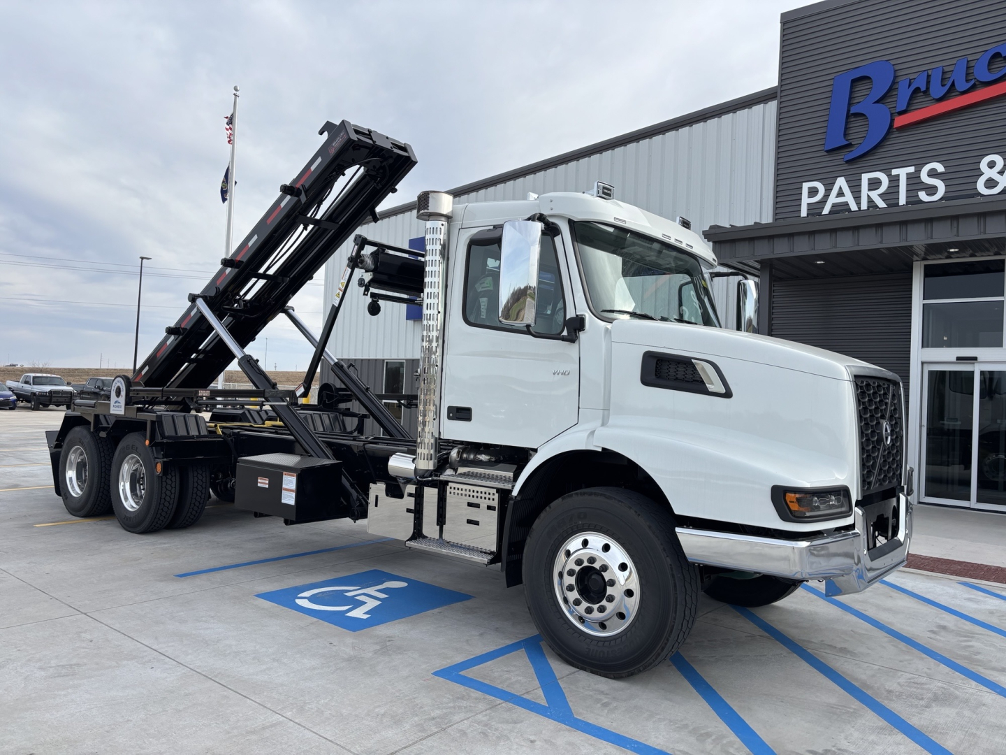 Volvo VHD64B300 roll-off truck in white cab with black body chassis, loading bed raised, parked in handicap space outside parts building.