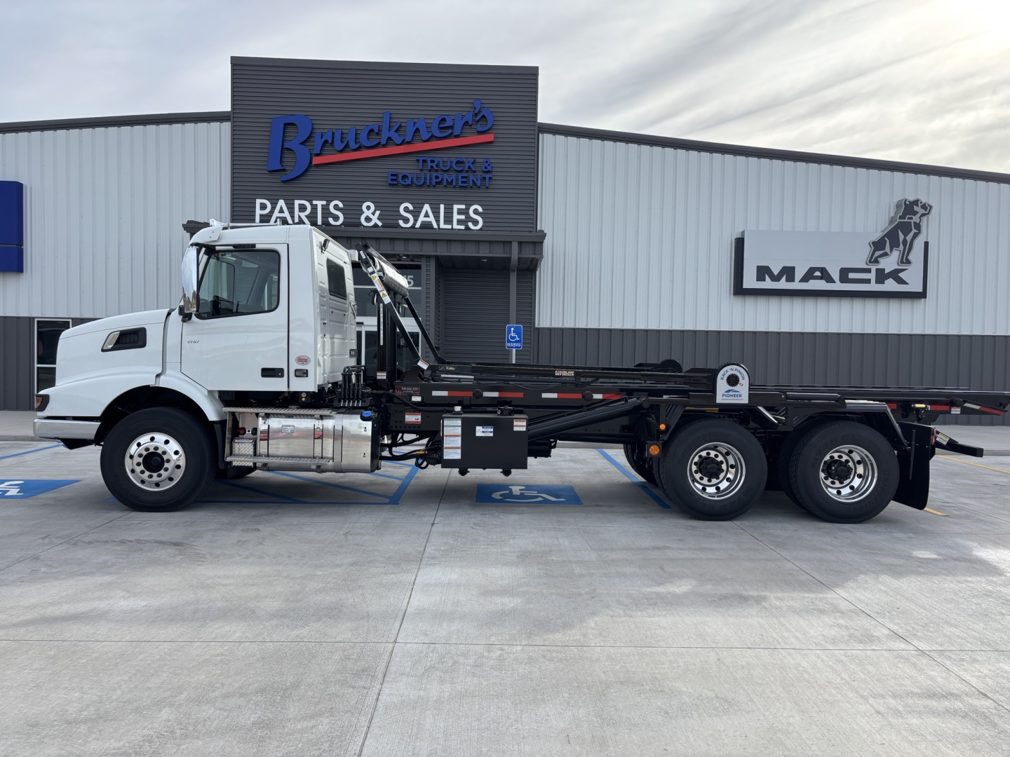 Volvo VHD64B300 roll-off truck side view showing cab, extended chassis, and flatbed with multiple axles in front of a parts and sales shop.