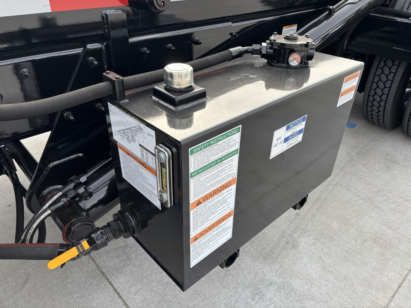 Truck chassis area showing a black rectangular tank with warning labels and caps; hoses and cables connected to left side; part of Volvo VHD64B300 truck.