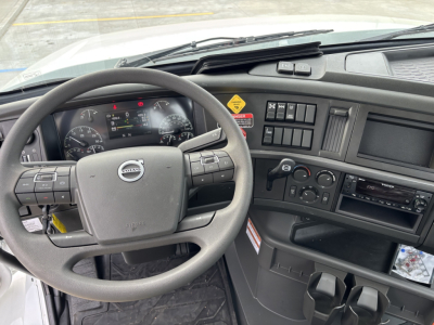 Volvo truck cockpit with steering wheel and dashboard, multiple control switches, circular gauges, and center console; visible labels include Volvo brand emblem on steering wheel.