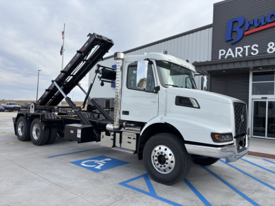 Volvo VHD64B300 roll-off truck in white cab with black body chassis, loading bed raised, parked in handicap space outside parts building.