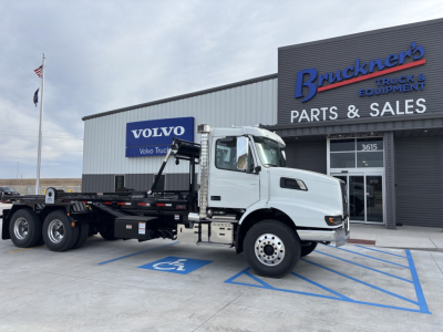 Volvo VHD64B300 roll-off truck parked in front of a dealership showing Volvo logo on building.