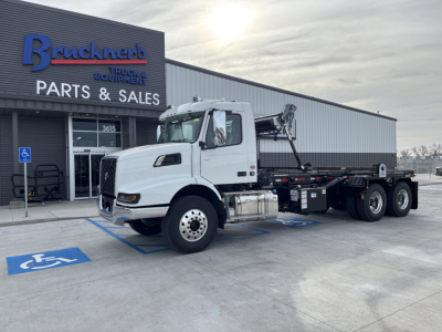 Volvo VHD64B300 rolloff truck in front of Bruckner's Parts &amp; Sales building showing white cab and hooklift bed.