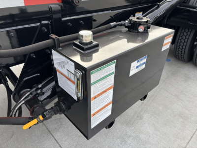 Truck chassis area showing a black rectangular tank with warning labels and caps; hoses and cables connected to left side; part of Volvo VHD64B300 truck.