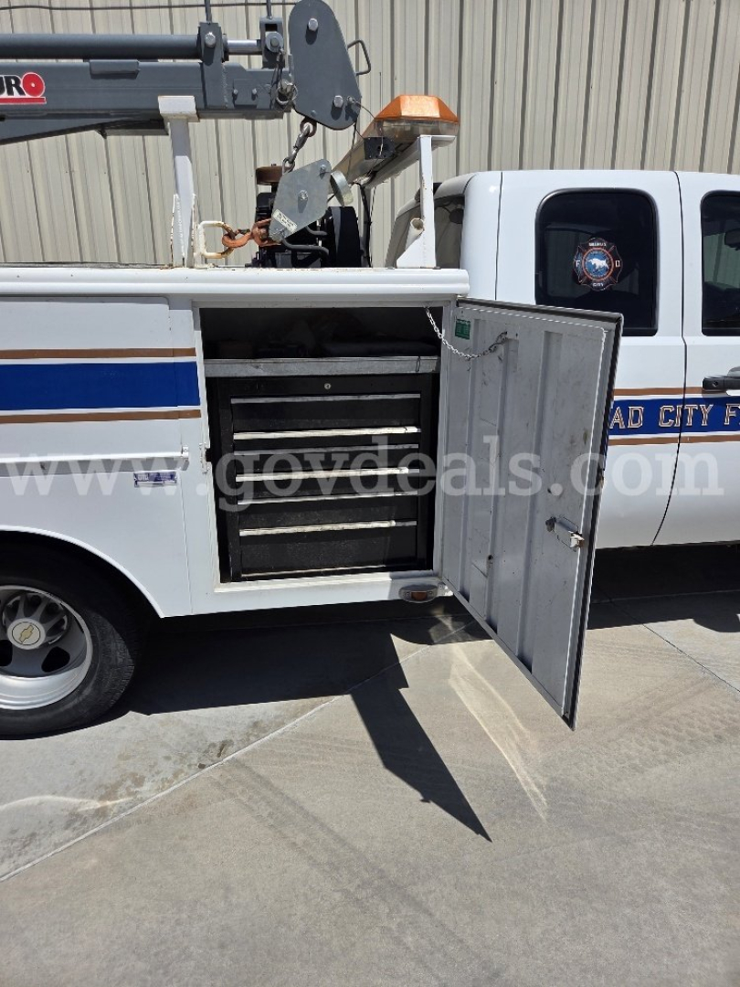 Truck service body with open side cabinet showing multiple drawers and a utility crane on top; white Chevrolet Silverado 3500HD pickup in background.