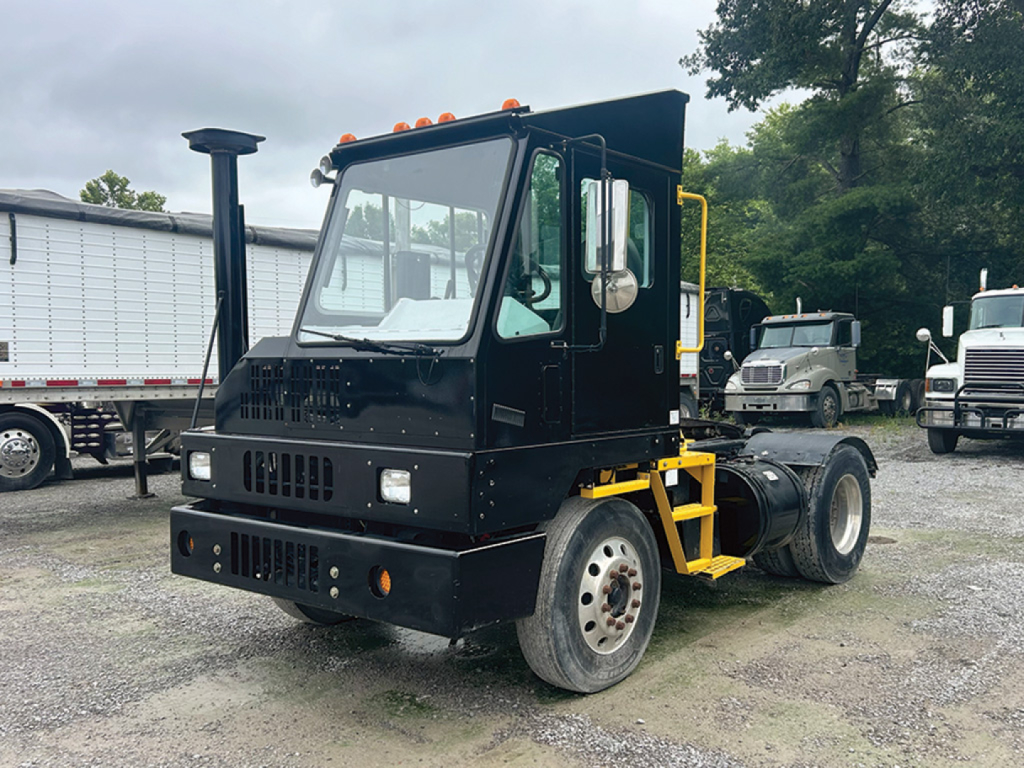 Kalmar Ottawa yard truck with raised roof cab and black paint, front view showing large rectangular grille, headlights, orange clearance lights, exhaust stack, and yellow rear frame with dual rear wheels.